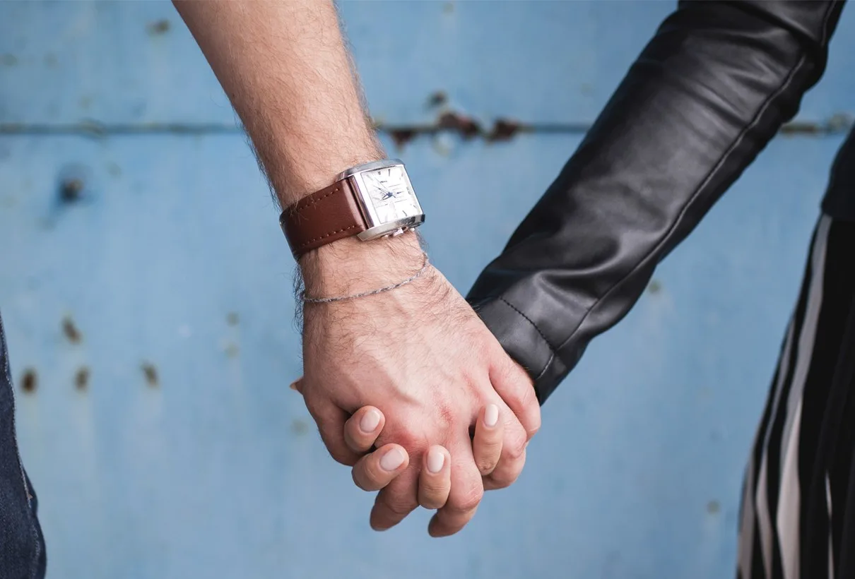 Två händer som håller varandra, den ena med armbandsur, mot en blå vägg