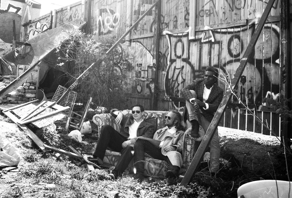 Svartvitt foto av fyra personer i kostym som sitter bland graffiti och bråte